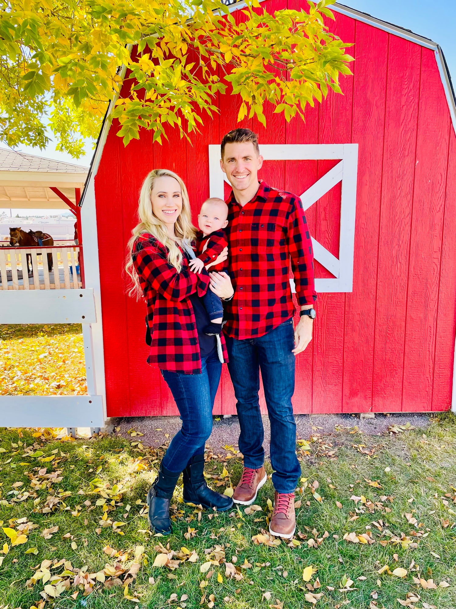 picture of michael and his wife and son during the fall season. standing in front of a barn with leaves changing and falling to the ground.